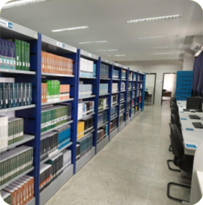 Foto mostrando um dos corredores da biblioteca. De um lado há estantes com livros e do outro há mesas com computadores