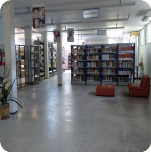 Imagem mostrando interior da biblioteca. Ao fundo aparecem estante com livros e sofás.