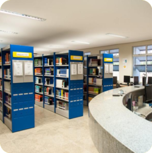 Foto mostrando estantes da biblioteca. A direta aparece parte do balcão de atendimento