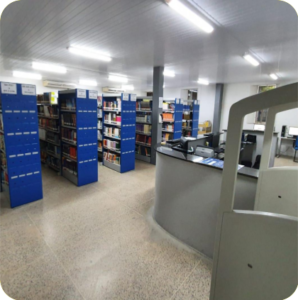 Imagem mostrando corredores de estantes de livros. Na direita aparece o balcão de atendimento da biblioteca.
