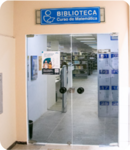 Fotografia colorida mostrando a entrada da BCM. A porta da biblioteca é de vidro e através dela é possível ver o interior da biblioteca, incluindo parte dos armários e do acervo.