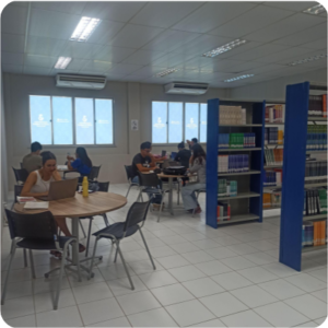 Fotografia colorida mostrando mesas circulares e cadeiras. Nas mesas há pessoas estudando. Na direita aparecem estantes azuis com livros.