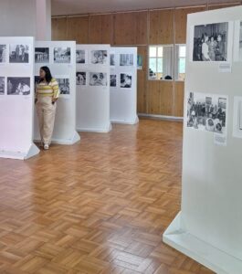 Foto colorida na vertical. Em um hall com chão de taco aparecem painéis brancos com fotos dispostas neles. No canto esquerdo aparece uma mulher, vestida de amarelo, olhando as fotos expostas.